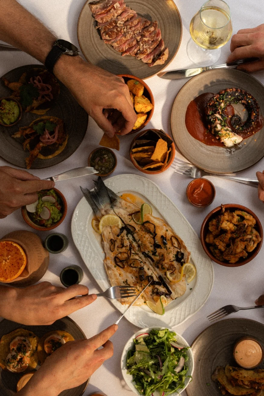 Overhead sharing feast at Pūblico Mykonos — whole grilled Aegean fish, octopus, steak, guacamole and totopos on white tablecloth